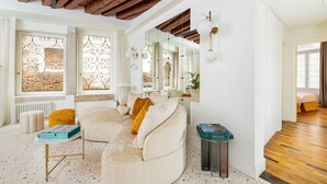 A modern living room with a large beige sectional sofa, a small round ottoman, a coffee table, and a side table with books. There are large windows with decorative iron grilles, a wooden ceiling, and a mirror reflecting the room. - Charming Canal View Apartment in Idyllic Location (Venecia)