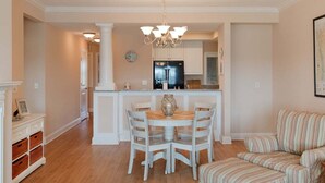 A dining area with a round table and chairs, a chandelier, and a kitchen in the background. - New! Condo with Dock + Pool (Carolina Beach)