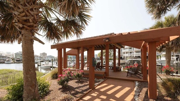A wooden deck with a pergola, surrounded by palm trees and flowering plants, overlooking a marina with boats. - New! Condo with Dock + Pool (Carolina Beach)
