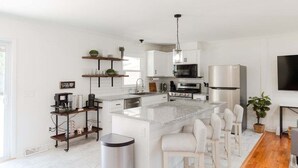 A modern kitchen with white cabinetry, a central island with bar stools, a stainless steel refrigerator, and a large window providing natural light. - New! Game Room + Close to Beach (Wilmington)