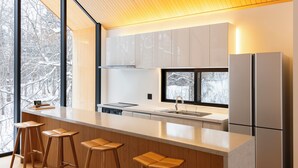 A modern kitchen with a large island, bar stools, stainless steel appliances, and a view of snow-covered trees. - Kouzan Woods (Niseko)
