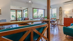 A spacious living area with a wooden bench, a dining table, and a sofa. There are large windows providing natural light and a view of the outdoors.