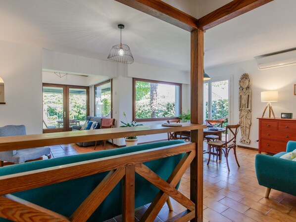 A spacious living area with a wooden bench, a dining table, and a sofa. There are large windows providing natural light and a view of the outdoors.