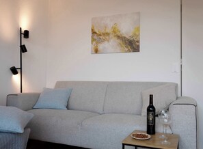 Un salon moderne avec un canapé gris, une petite table sur laquelle se trouvent une bouteille et des verres, et un tableau abstrait accroché au mur.