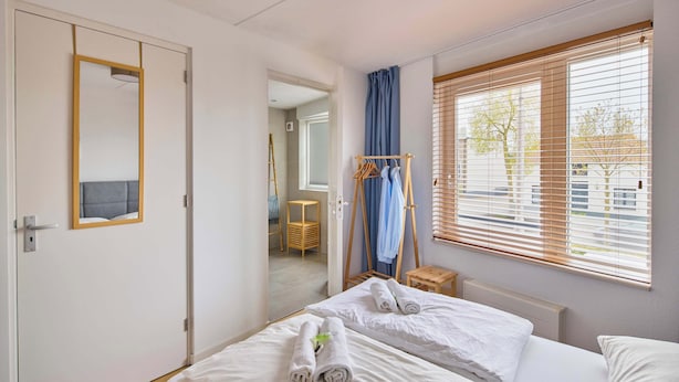 A hotel room with a bed, a mirror, a window with blinds, a hanging clothes rack, and a small stool. - Nimma Voorstad Stay Nijmegen (Nijmegen)