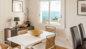 A dining area with a white table, wicker chairs, and a large bowl centerpiece. A window offers a view of the sea.