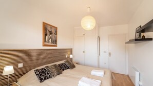 A modern bedroom with a wooden headboard, a large bed with patterned pillows, a bedside table with a lamp, and a hanging light fixture.