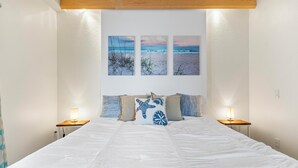 Un dormitorio con una cama grande, dos mesitas de noche con lámparas, y tres cuadros colgados en la pared que muestran escenas de playa.