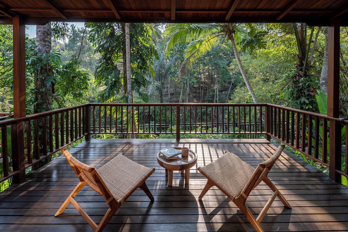 A wooden deck with two wicker chairs and a small table, surrounded by lush tropical vegetation.