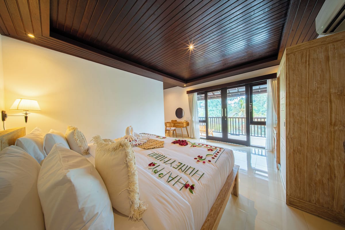 A spacious bedroom with a large bed, wooden ceiling, and a view of the outdoors through a sliding door.