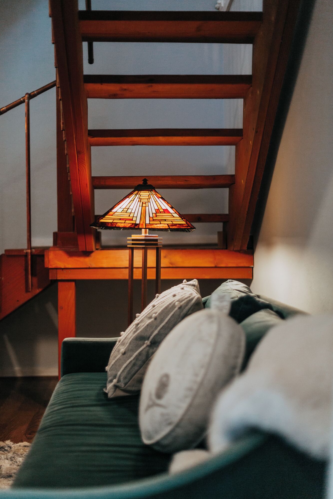 A room with a wooden staircase, a green sofa with pillows, and a table with a stained glass lamp.