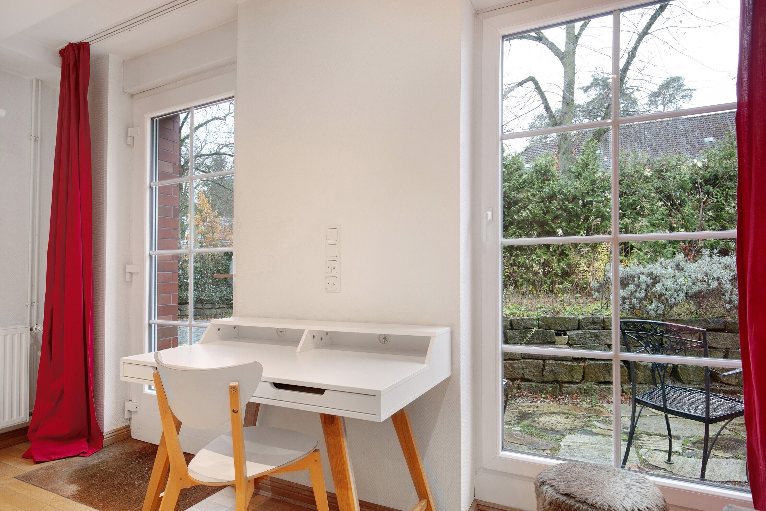 A room with a white desk, two chairs, a window with a view of a garden, and a red curtain.