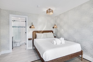 Une chambre Ă coucher avec un lit en bois, du linge de lit blanc, une salle de bain avec des toilettes et une douche, et un papier peint Ă motifs.