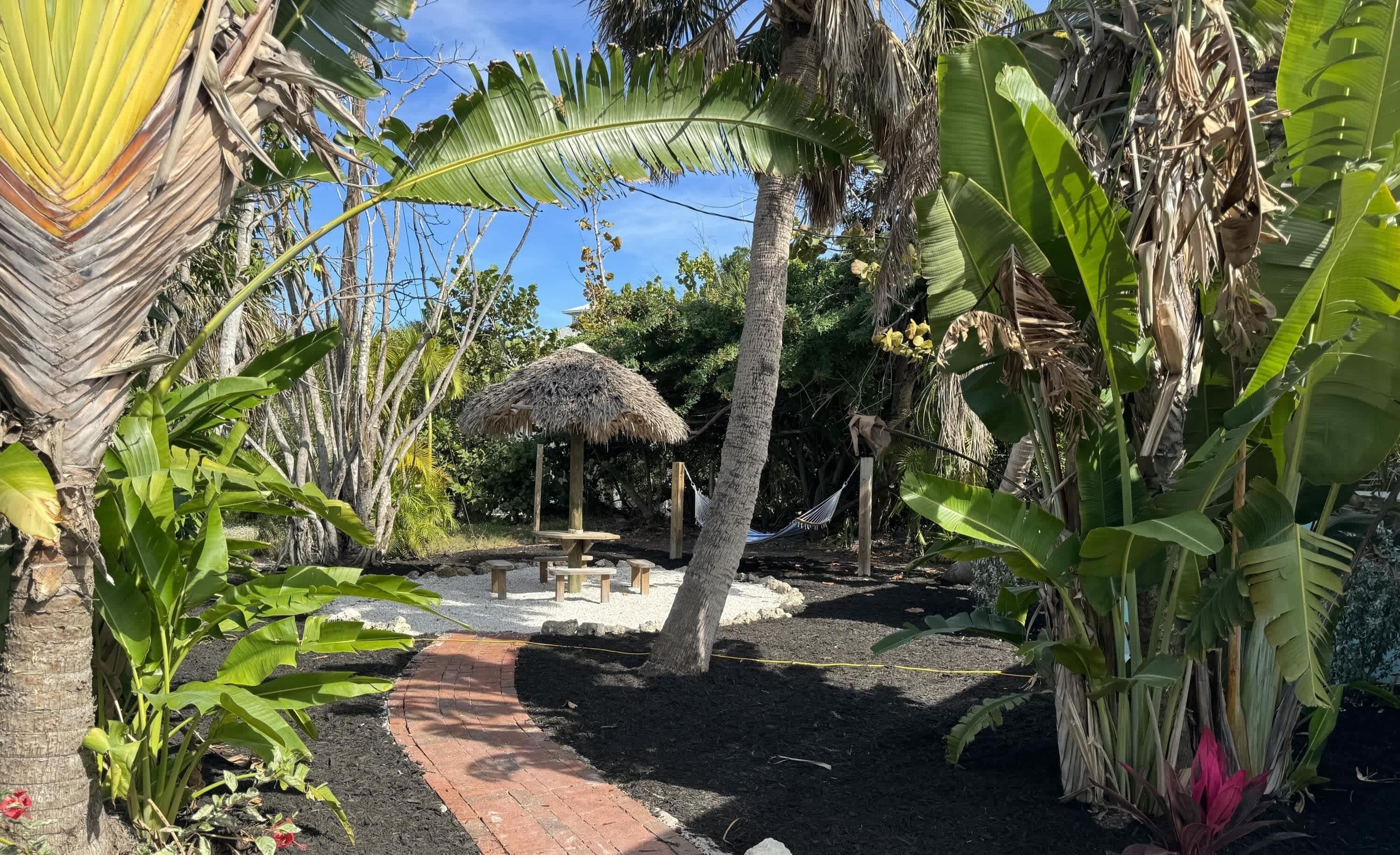 A garden with a thatched-roof structure, a brick pathway, and various tropical plants.