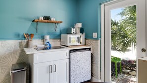 A compact kitchen with a microwave, toaster, and coffee maker on the counter, a small refrigerator, and a shelf with kitchen utensils. - The Palm Frond | Studio Cottage at Palmview of Sanibel with Bikes (Sanibel)
