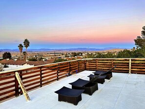 Une terrasse sur le toit offrant une vue sur le paysage urbain et les montagnes, agrémentée d’une clôture en bois et de plusieurs fauteuils de jardin en osier foncé.