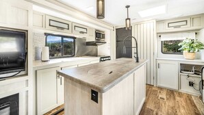 A modern RV kitchen with white cabinetry, a central island, a flat-screen TV, a microwave, and a sink. - Desert Tiny Home B25: Relaxing Desert Accommodation w/ Comfort Amenities (Jacumba Hot Springs)