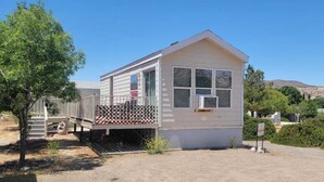 Une maison mobile de plain-pied, dotée d’un porche à l’avant et d’une petite terrasse, entourée d’arbres et sous un ciel dégagé.