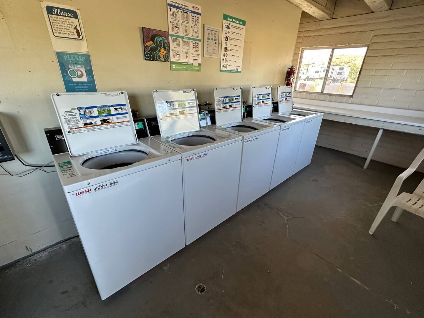 Une buanderie équipée de quatre machines à laver et de quatre sèche-linge, avec des affiches explicatives accrochées au mur et une fenêtre laissant entrer la lumière naturelle.