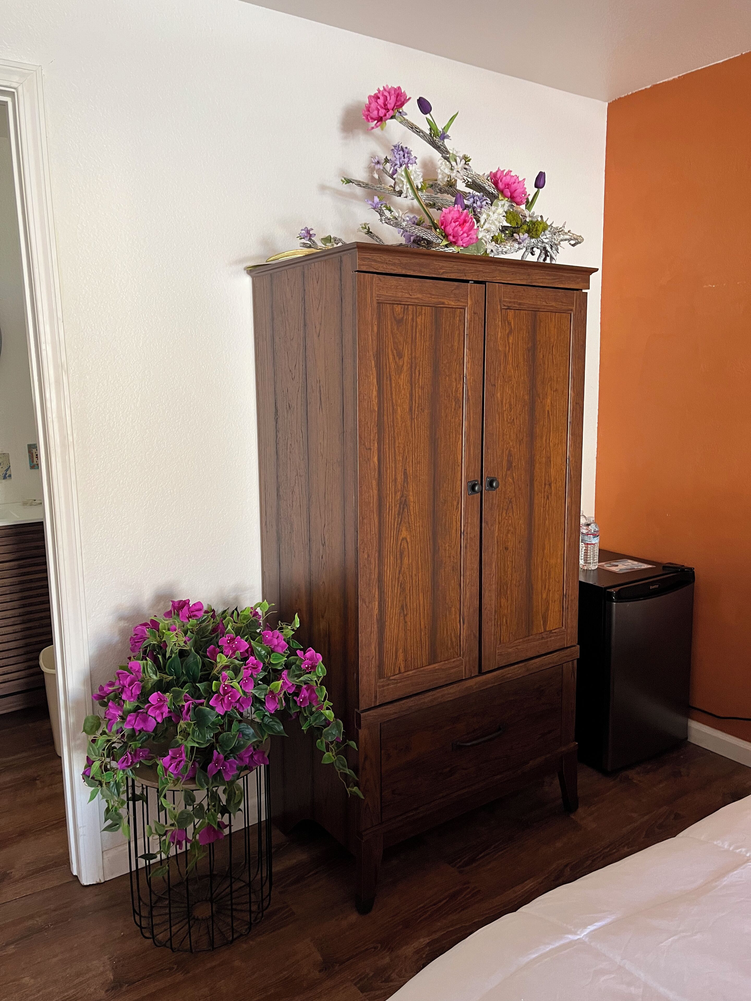 Un armario de madera con dos puertas, una mesita con una botella de agua y una maceta con flores moradas.