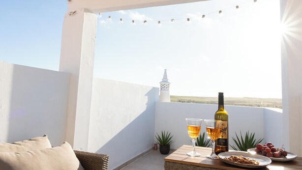 Ein Balkon mit einer Rattan-Couch, einem Tisch mit einer Weinflasche, Gläsern und einer Traubenteller.
