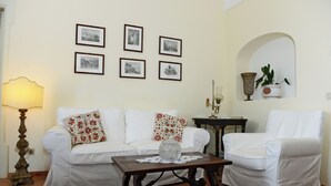 A living room with a white sofa, a matching armchair, a wooden coffee table, and a patterned rug. There are four framed pictures on the wall and a lamp on the floor. - Ancient mansion with breathtaking views amid ancient olive and citrus trees (Praiano)