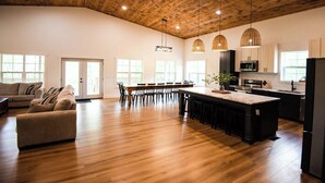 Un salón amplio con techo de madera, una mesa de comedor con sillas, una zona de cocina con electrodomésticos modernos y un gran ventanal que proporciona luz natural.