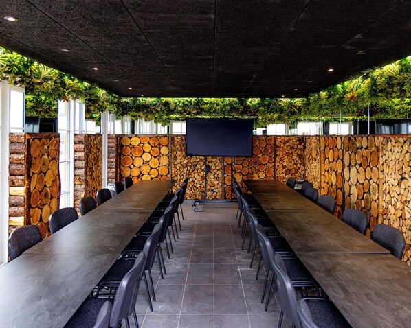 A conference room with a long table, chairs, a screen, and a wall decorated with logs and greenery. - Fun Art Blokhus (Blokhus)