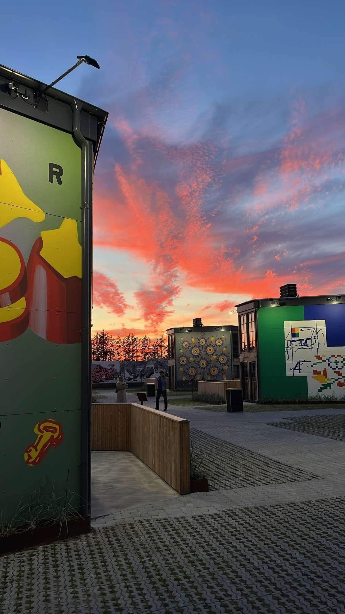 Un bâtiment à la façade verte et surmonté d’une enseigne jaune en forme de flèche, une zone pavée avec un banc en bois, et un ciel au coucher du soleil.
