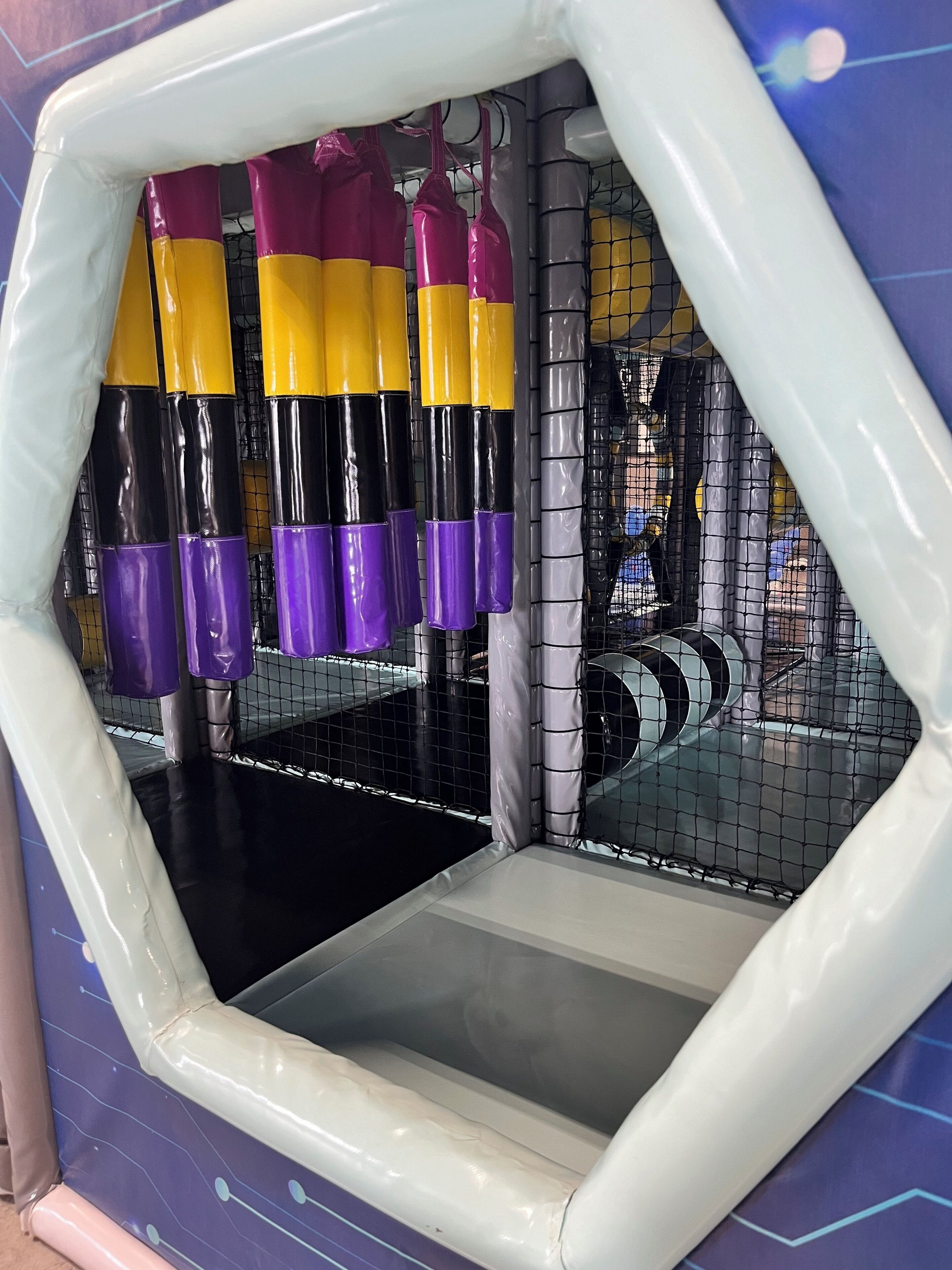 An indoor play area with a padded floor, netted walls, and colorful hanging toys.