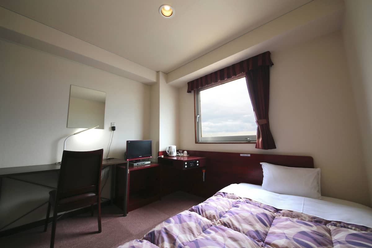 A hotel room with a bed, a desk with a computer, a chair, a window with curtains, and a ceiling light.