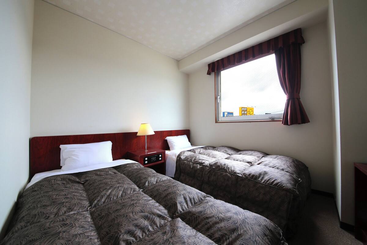 A hotel room with two beds, a nightstand, a lamp, and a window with curtains.