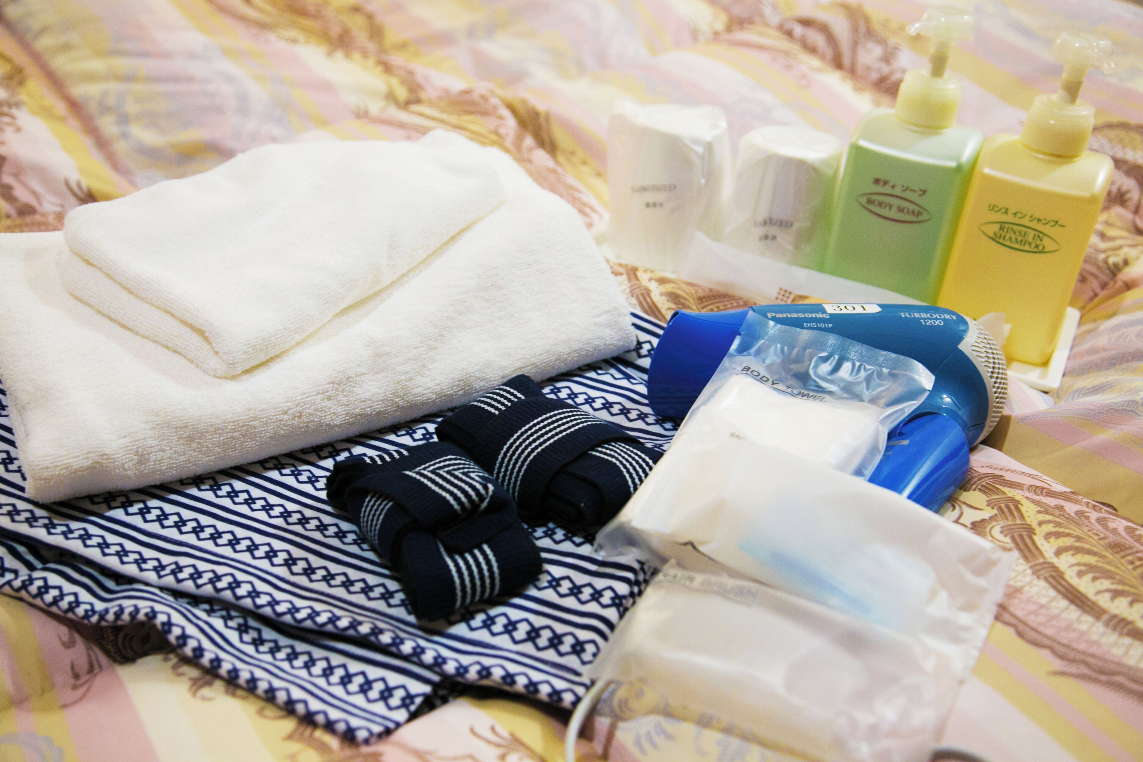 A neatly arranged bed with a folded white towel, a patterned cloth, and toiletries including body wash, shampoo, and a hairdryer.
