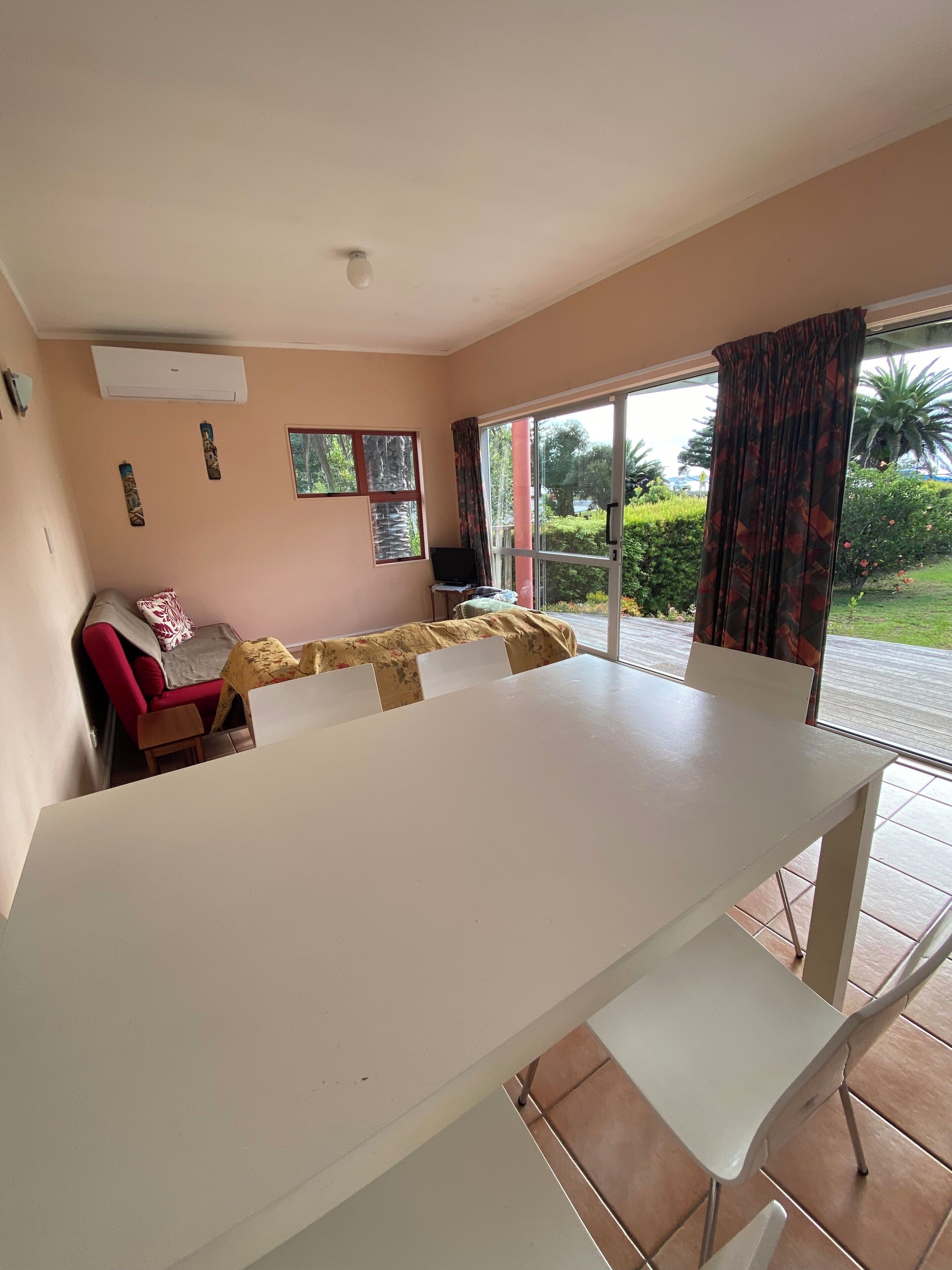 A room with a large white table, a red sofa, and a laptop on a small table. There is a sliding glass door leading to an outdoor area with greenery.