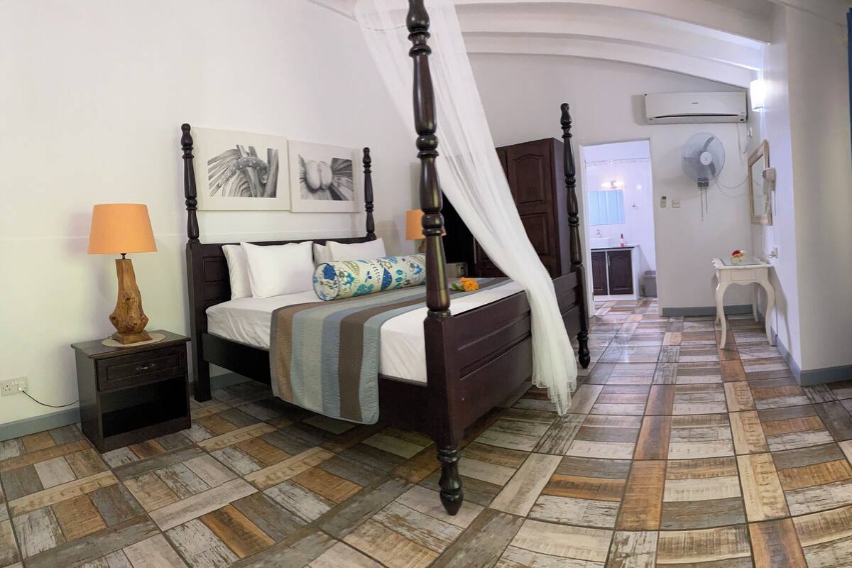 A bedroom with a four-poster bed, a bedside table with a lamp, a bedside plant, a large window with curtains, and a fan on the wall.