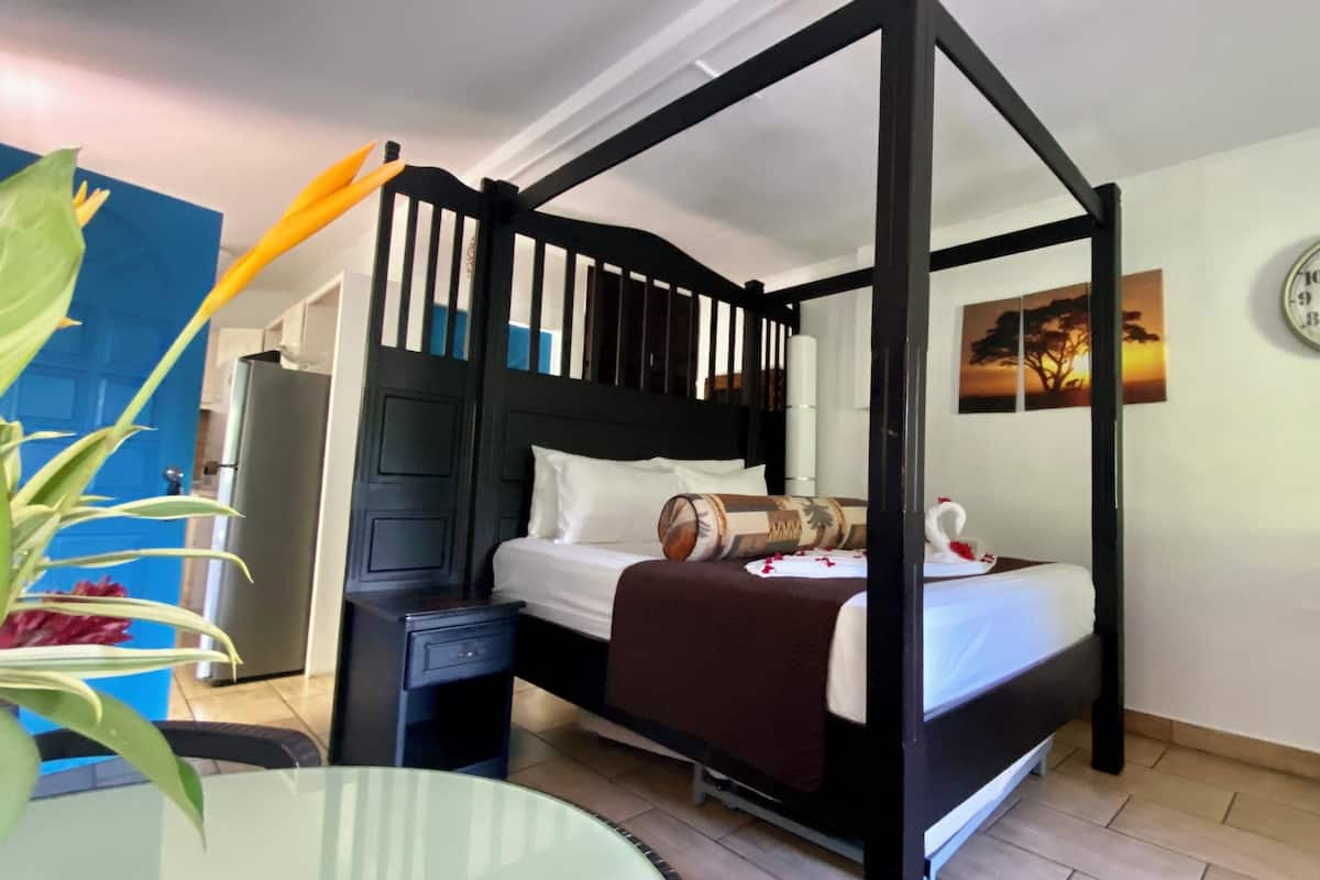A four-poster bed with a dark headboard, a bedside table, a clock, and a wall painting.