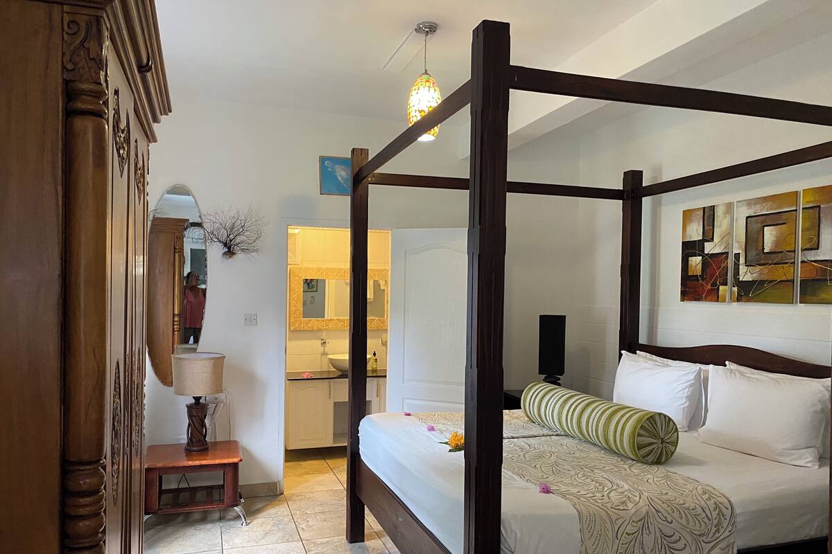 A four-poster bed with a white bedspread and green striped pillow, a wooden nightstand with a lamp, and a bathroom with a mirror and sink.
