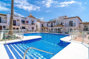 A swimming pool surrounded by white buildings with balconies, a clear blue sky, and a palm tree.