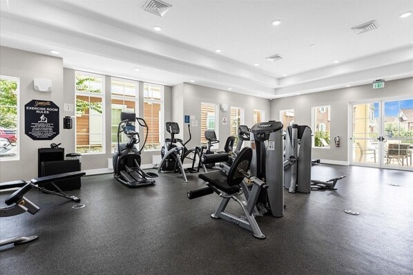 Ein gut beleuchteter Fitnessraum mit verschiedenen Trainingsgeräten, großen Fenstern und einem Schild mit den Regeln für den Trainingsraum.