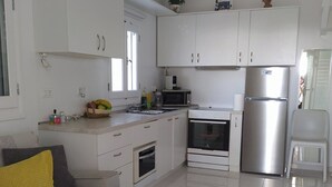 A modern kitchen with white cabinets, a stainless steel refrigerator, a microwave, and an oven. There is a small dining area with a white chair and a sofa with a yellow pillow.