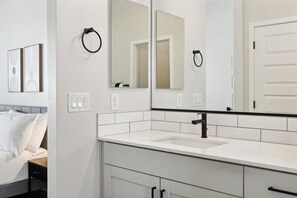 A modern bathroom with a white sink, black faucet, and a large mirror. There are two towel rings on the wall and a white door. - Sparkling Modern Home in Canopy w/ Pool,BBQ (Buckeye)