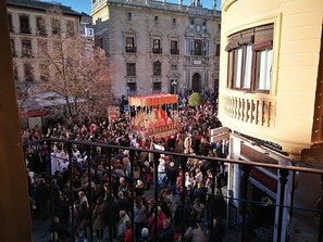 Une place publique animée où une foule s’est rassemblée autour d’un événement central, avec des bâtiments historiques en arrière-plan et un escalier menant à un niveau supérieur.