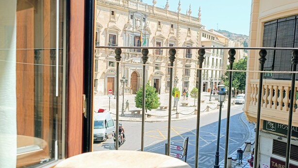Vue depuis un balcon, avec une table ronde et une rambarde en métal. Au fond, on aperçoit une rue bordée d’immeubles, un beffroi et un fourgon blanc.