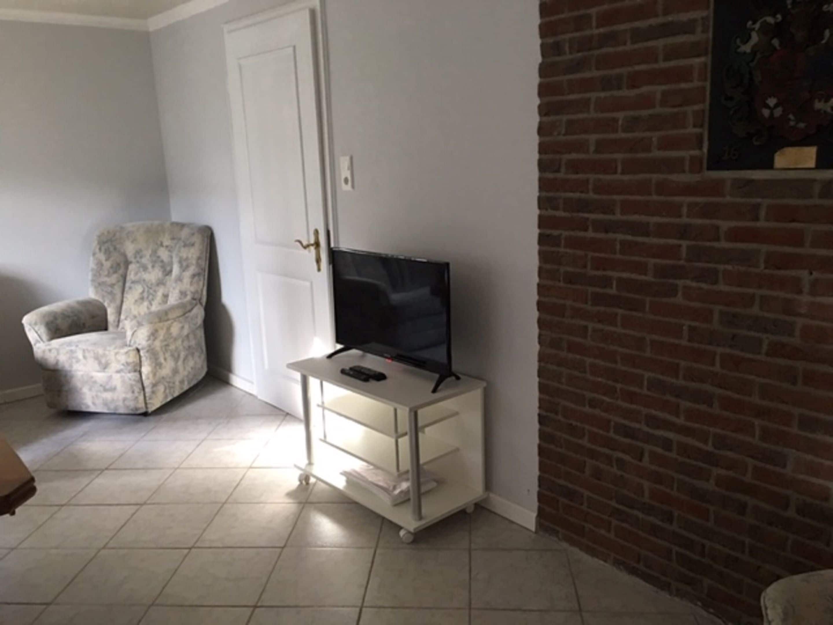 Una habitación con una puerta blanca, un televisor sobre un pedestal blanco, un sillón estampado y una pared de ladrillo.