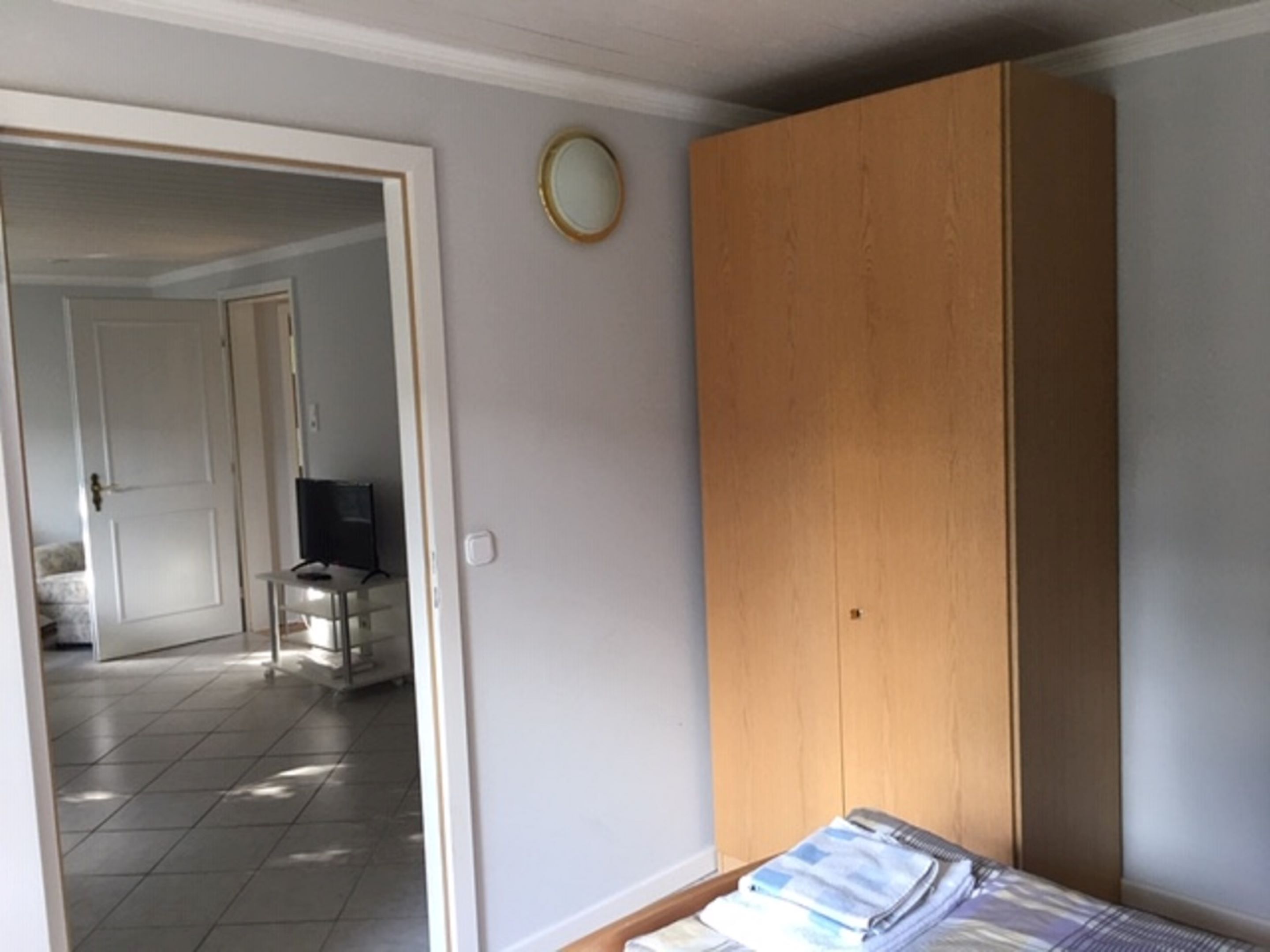 A bedroom with a wooden wardrobe, a bed with a blue and white striped bedspread, and a small round mirror on the wall.