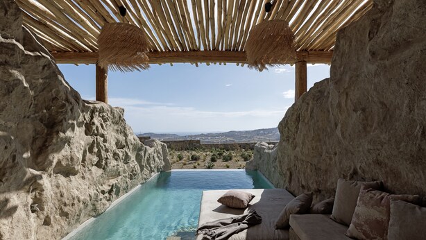 A stone-walled infinity pool with a wooden pergola overhead, a beige sofa with cushions, and a view of distant hills. - WhiteArt Suites (Mykonos)