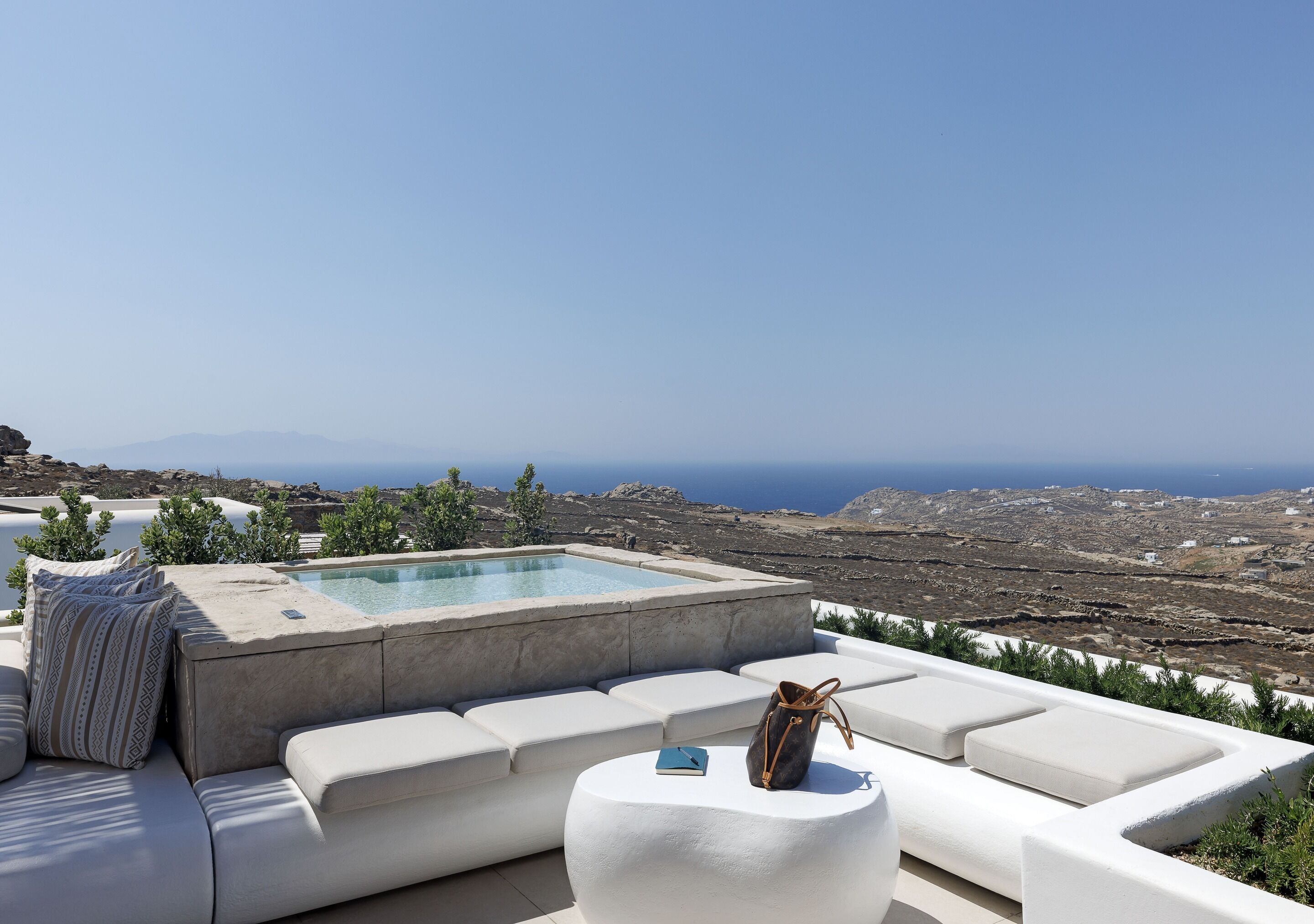 A poolside area with a hot tub, white cushioned seating, and a view of the sea and mountains.