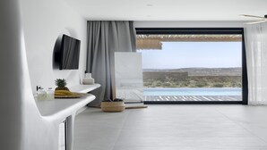 A modern hotel room with a large window, a flat-screen TV, a bed, and a small table with a pineapple and bananas. - WhiteArt Suites (Mykonos)