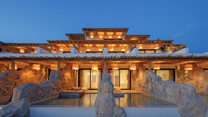 A multi-story building with a pool in the foreground, large rock formations, and warm lighting. - WhiteArt Suites (Mykonos)