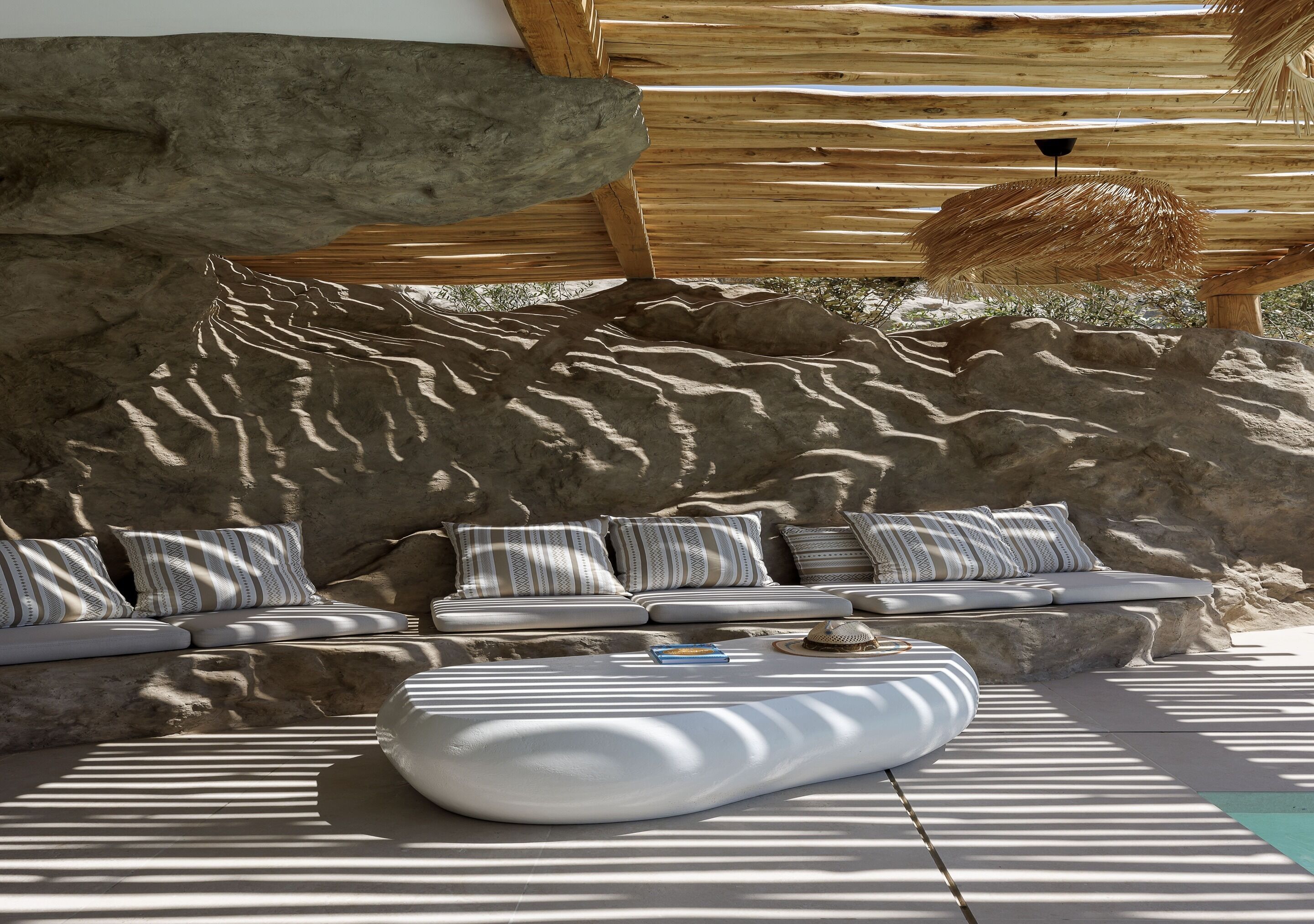 A sunlit outdoor seating area with a stone wall backdrop, cushioned seating, and a woven umbrella.